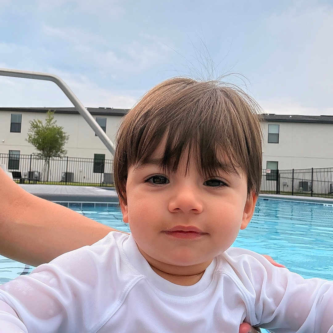 Isaac is registered to the contest to win money with this photo: toddler, child, swimming_pool, water, white_shirt, hand, building, sky, outdoor, residential, fence, support, portrait, cute, young_child, person, face, hair, expression, daytime