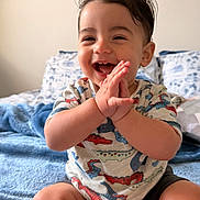 Isaac is registered to the contest to win money with this photo: baby, child, smiling, clapping, dinosaur_print, shirt, blue_blanket, bed, pillow, indoor, cute, happy, sitting, toes, hands, young_child, portrait, room, cozy, casual