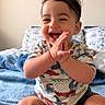 baby, child, smiling, clapping, dinosaur_print, shirt, blue_blanket, bed, pillow, indoor, cute, happy, sitting, toes, hands, young_child, portrait, room, cozy, casual