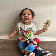 Mateo is registered to the contest to win money with this photo: child, toddler, smiling, happy, sitting, portrait, indoor, home, floor, wall, wood_floor, toy, plush_toy, super_mario, paddle, t_shirt, jeans, sneakers, curly_hair, playtime