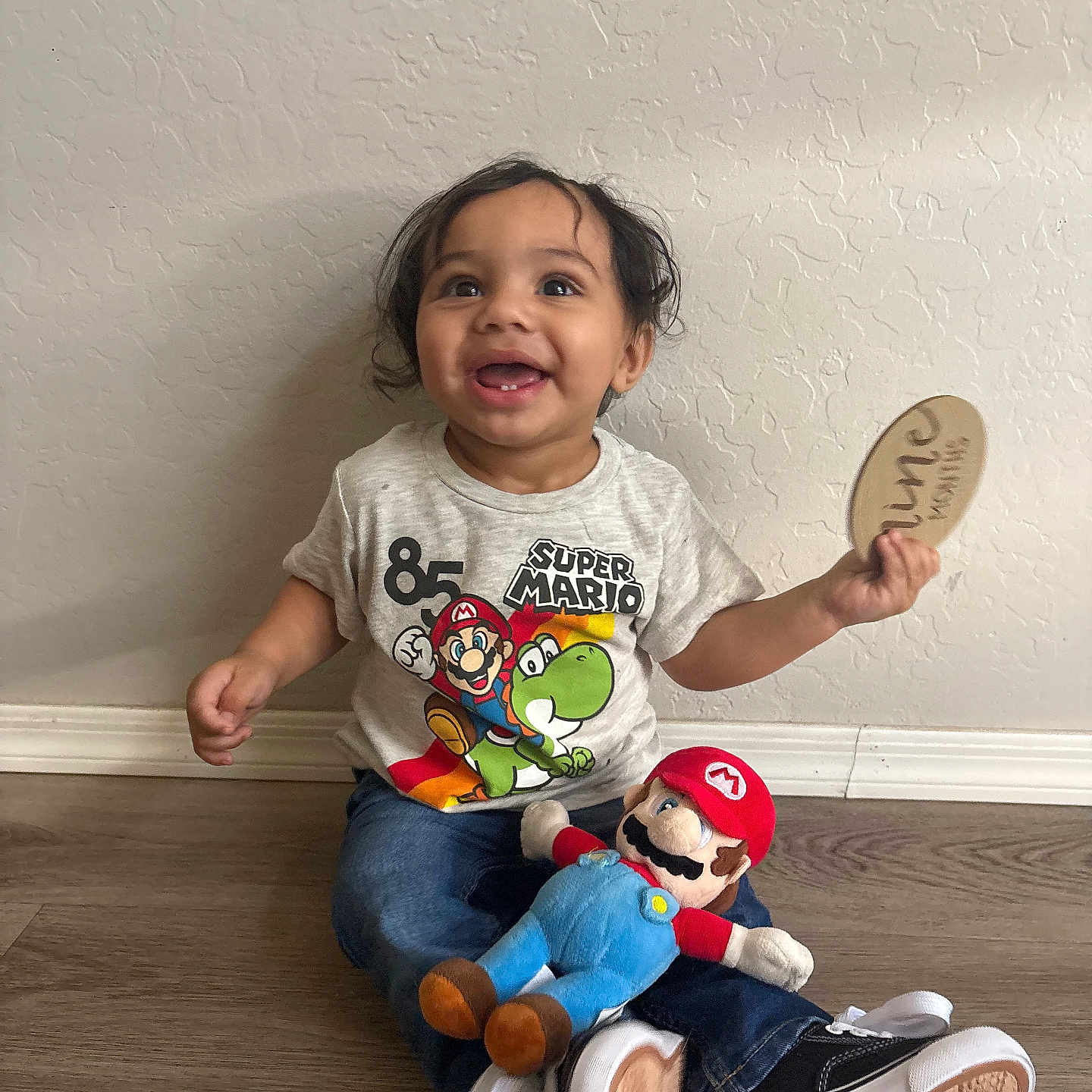 Mateo is registered to the contest to win money with this photo: child, toddler, smiling, happy, sitting, portrait, indoor, home, floor, wall, wood_floor, toy, plush_toy, super_mario, paddle, t_shirt, jeans, sneakers, curly_hair, playtime