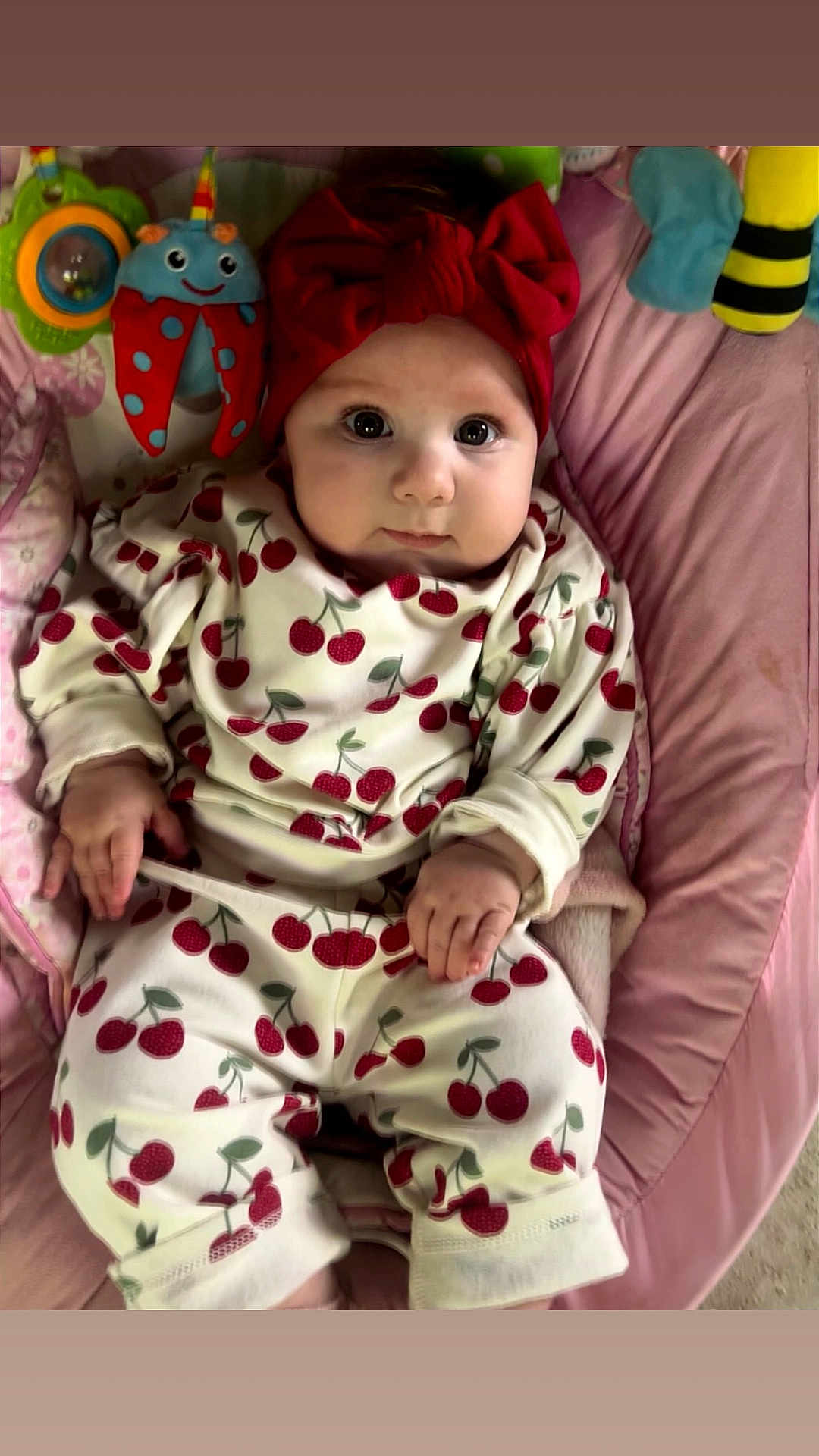 Marlie is registered to the contest to win money with this photo: baby, infant, child, red_bow, cherry_pattern, clothing, pink_cushion, toy, colorful_toy, face, hand, lying_down, cute, wide_eyes, headband, comfort, indoor, portrait, baby_outfit, soft_fabric