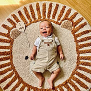 Côme a rejoint le concours — aidez-le/la à gagner de superbes lots ! baby, infant, smiling, lying_down, rug, floor, wooden_floor, lion_design, plush_rug, clothing, overalls, white_shirt, wooden_sign, text, sunlight, shadow, happy, child, indoors, cute