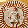 baby, infant, smiling, lying_down, rug, floor, wooden_floor, lion_design, plush_rug, clothing, overalls, white_shirt, wooden_sign, text, sunlight, shadow, happy, child, indoors, cute