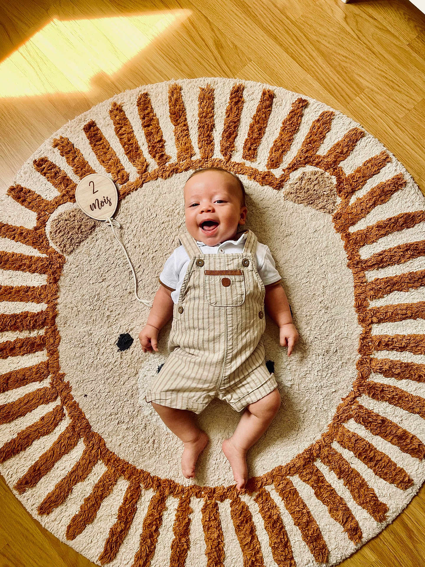 Côme a rejoint le concours — aidez-le/la à gagner de superbes lots ! baby, infant, smiling, lying_down, rug, floor, wooden_floor, lion_design, plush_rug, clothing, overalls, white_shirt, wooden_sign, text, sunlight, shadow, happy, child, indoors, cute