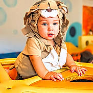 Max is registered to the contest to win money with this photo: toddler, child, costume, lion_costume, indoor, play_area, yellow, curious, baby, cute, portrait, face, hands, soft_toys, playground, young_child, clothing, expression, headwear, colorful_background