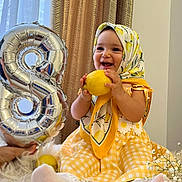 Zina a rejoint le concours — aidez-le/la à gagner de superbes lots ! baby, child, toddler, smiling, yellow_dress, headscarf, lemon, balloon, number_8, curtains, flowers, indoor, happy, celebration, holding, sitting, cute, portrait, bright, decor