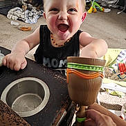 Vincent is registered to the contest to win money with this photo: toddler, child, happy, smiling, toy, goblet, hand, table, living_room, carpet, dog, dog_crate, blanket, furniture, person, indoor, playful, expression, home, baby