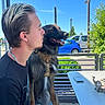bench, blue_sky, casual_clothing, chain_necklace, daylight, dog, greenery, happy_dog, harness, man, muzzle, outdoor, parked_cars, pet, phone, relaxed, short_hair, side_profile, sunny, table
