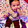 baby, bow, child, colorful, cushion, cute, earrings, eyes, face, floral_pattern, hand, headband, indoors, knitted_clothing, person, playful, sitting, smiling, string, toy_phone