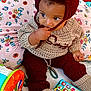 Saniyah is registered to the contest to win money with this photo: baby, beige, blanket, chewing, child, colorful, couch, curious, cute, floral, footwear, hand, hat, indoor, knitwear, maroon, pillow, play_phone, sitting, toy