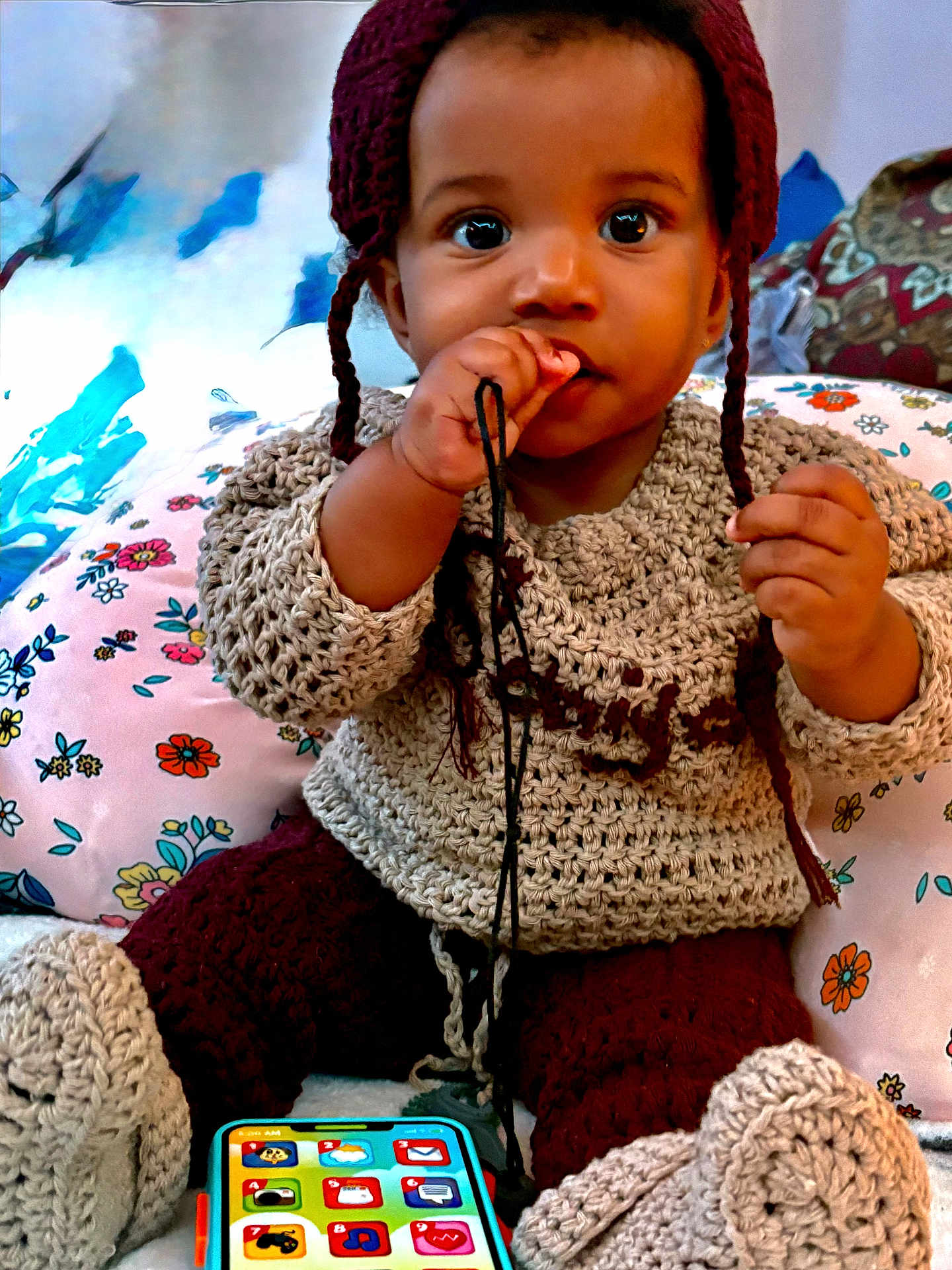 Saniyah joined the competition — help win amazing prizes! baby, child, closeup, cozy, curious, cute, floral_pattern, handmade, hat, indoor, infant, knitted_clothing, pillow, playful, portrait, sitting, smiling, string, toy_phone, warm_colors