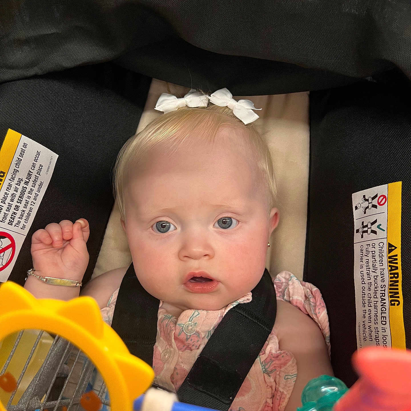 Angelica is registered to the contest to win money with this photo: accessory, baby, blond_hair, blue_eyes, bracelet, car_seat, child, closeup, curious, face, indoors, infant, pink_dress, playful, portrait, seatbelt, sitting, straps, toy, white_bow
