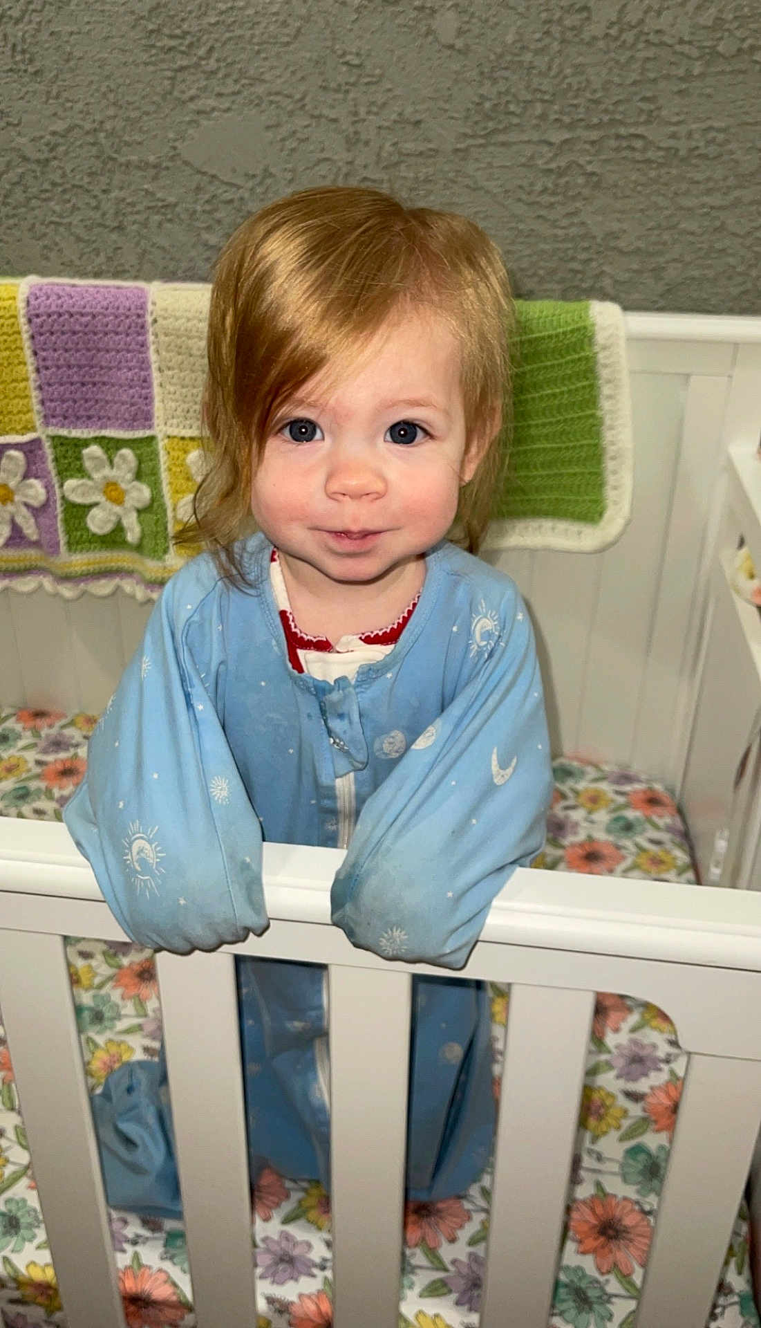 Bennett is registered to the contest to win money with this photo: child, toddler, crib, pajamas, blanket, crochet, nursery, portrait, smile, blue_pajamas, standing, hands_on_rail, floral_bedding, hair, eyes, face, indoor, wooden_crib, baby, cozy