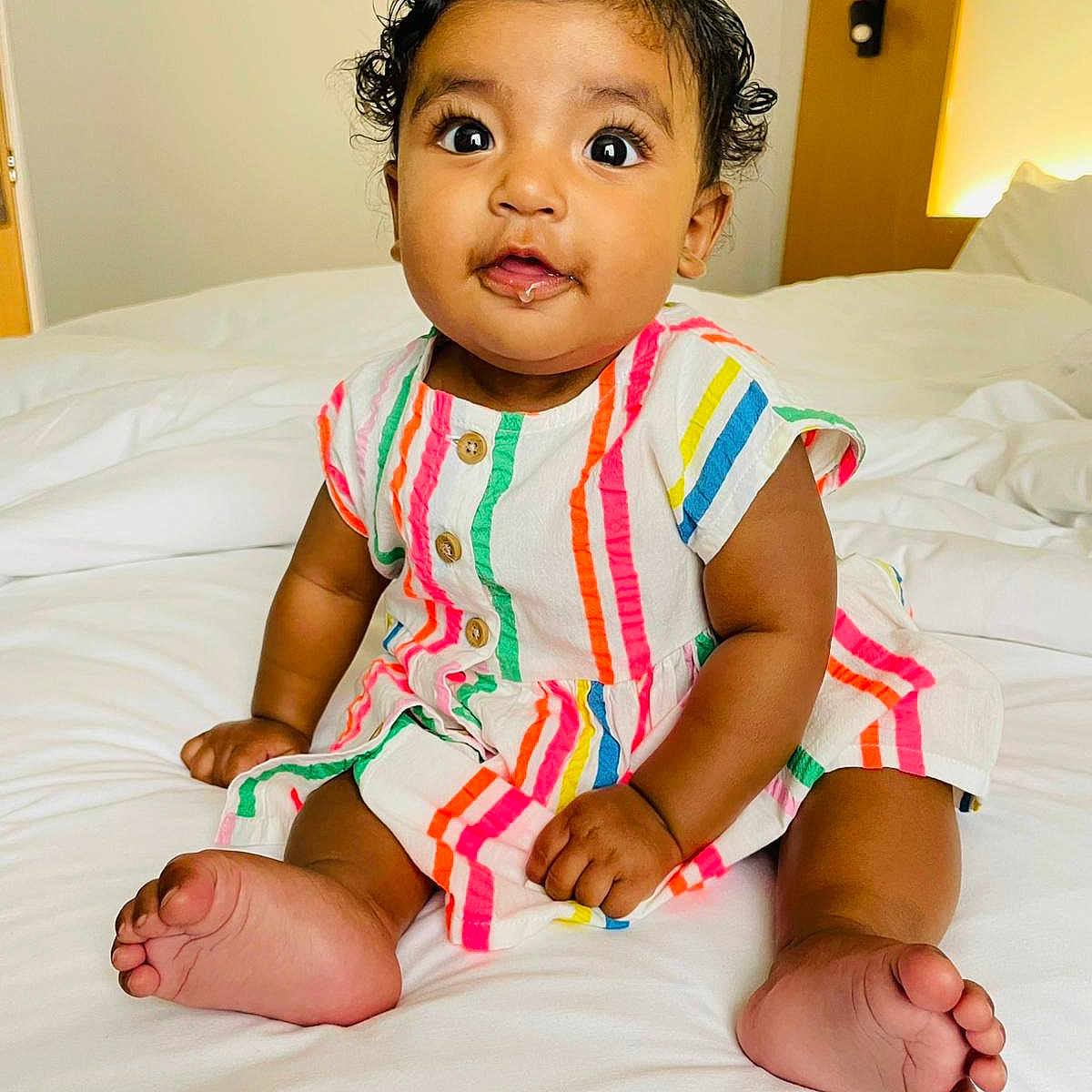 Sierra is registered to the contest to win money with this photo: baby, bed, child, curious, cute, drool, foot, hand, happy, indoor, infant, person, pink_hair_clip, portrait, room, sitting, skin, soft_lighting, striped_dress, white_bedding