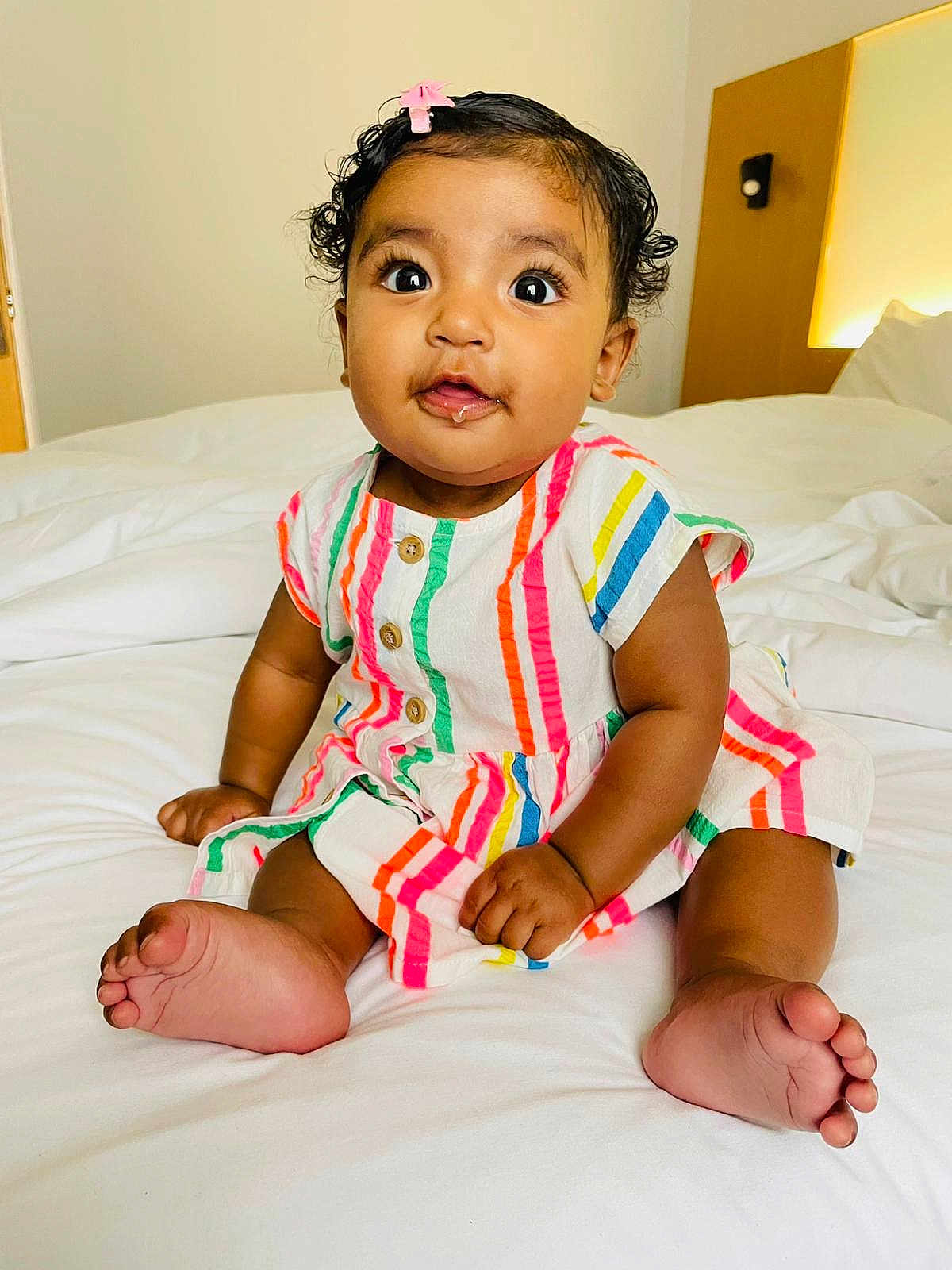 Sierra is registered to the contest to win money with this photo: baby, bed, child, curious, cute, drool, foot, hand, happy, indoor, infant, person, pink_hair_clip, portrait, room, sitting, skin, soft_lighting, striped_dress, white_bedding