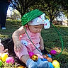 baby, child, grass, easter_eggs, basket, headband, pink_clothing, socks, outdoor, trees, sunlight, person, toy, colorful, nature, playful, cute, sitting, park, fun
