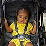 baby, child, stroller, yellow_outfit, harness, infant, seat, straps, curious, wide_eyes, indoor, padded, black, clothing, sitting, hands, legs, face, person, baby_girl