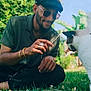 man, cat, grass, outdoor, greenery, sunlight, sunglasses, cap, beard, casual_clothing, nature, pet, happy, smiling, jewelry, bracelet, ring, playing, interaction, relaxation