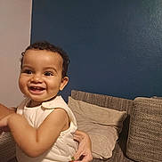 Ethan a rejoint le concours — aidez-le/la à gagner de superbes lots ! toddler, child, smile, happy, face, person, couch, pillow, hand, indoor, clothing, wall, blue_wall, beige_wall, texture, curly_hair, skin, sleeveless, holding, baby