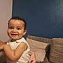 toddler, child, smile, happy, face, person, couch, pillow, hand, indoor, clothing, wall, blue_wall, beige_wall, texture, curly_hair, skin, sleeveless, holding, baby