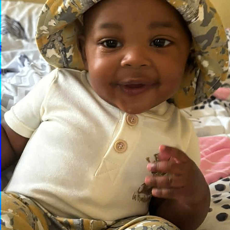 Jacyon is registered to the contest to win money with this photo: baby, child, infant, smiling, portrait, camouflage, hat, clothing, bed, bedroom, sitting, face, eyes, hands, indoors, cozy, fashion, cute, photography, happy