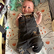 Jasper is registered to the contest to win money with this photo: baby, infant, child, overalls, white_socks, toys, dinosaur_mat, play_area, cute, smiling, indoor, soft_toys, baby_clothes, lying_down, floor, wooden_floor, happy, face, person, portrait