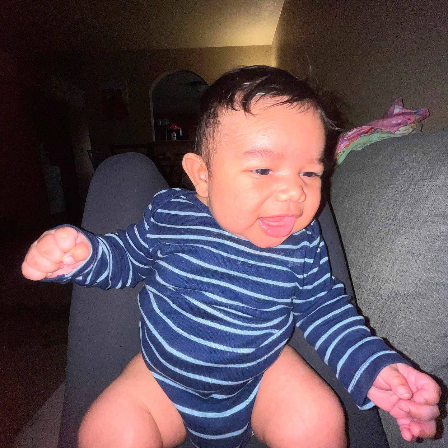Mateo is registered to the contest to win money with this photo: baby, infant, child, striped_onesie, blue_clothing, sitting, reaching, smiling, tongue_out, portrait, face, indoors, living_room, couch, chair, household, flash_photography, dark_background, colorful_blanket, happy