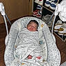 newborn, baby, infant, sleeping, bouncer, blanket, swaddled, face, portrait, indoors, sneakers, shoe_rack, wood_floor, furniture, nursery, cozy, relaxed, patterned_fabric, feet, photograph