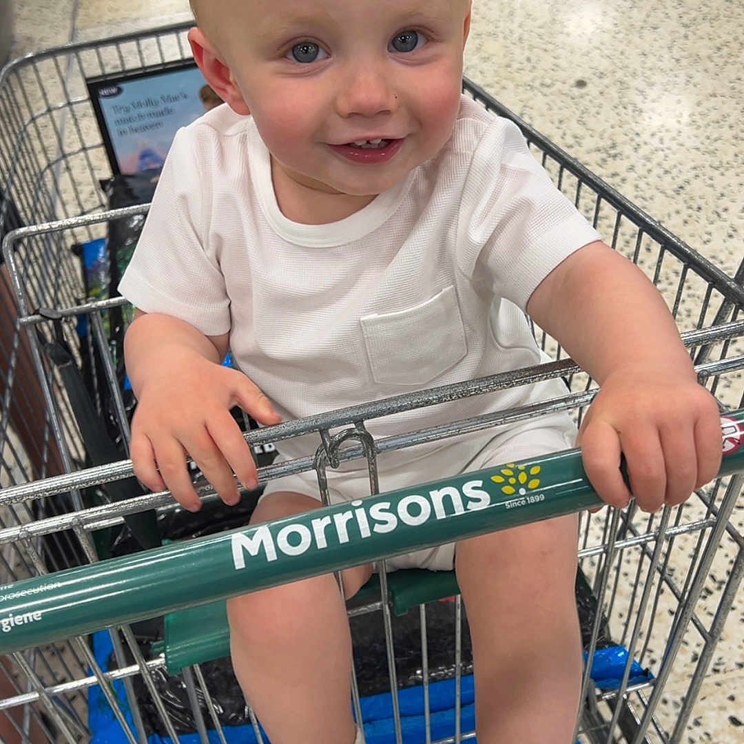 Reggie is registered to the contest to win money with this photo: blonde_hair, blue_eyes, child, floor, footwear, handlebar, happy, holding, indoor, metal, person, retail, shopping, shopping_cart, smiling, sneakers, store, supermarket, toddler, white_clothing