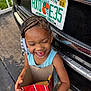 Kaylani is registered to the contest to win money with this photo: child, smiling, braided_hair, beads, candy, cardboard_box, vehicle, license_plate, outdoor, concrete, happy, person, tank_top, black_car, sunshine_state, florida, road, sidewalk, holding, portrait