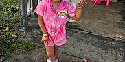 Kaylani is registered to the contest to win money with this photo: child, girl, pink_outfit, rainbow_patch, smiling, braided_hair, bracelets, sneakers, balloons, party, outdoor, concrete_floor, brick_path, fence, table, cake, chairs, people, sunlight, celebration
