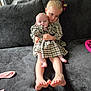 baby, child, sibling, couch, plaid_dress, indoor, family, hug, cute, blond_hair, foot, smile, person, home, toddler, hair_bow, seated, cozy, affection, portrait