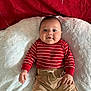 baby, infant, smile, red_striped_sweater, beige_shorts, blanket, soft_texture, face, child, cute, lying_down, indoors, skin, hand, head, eyebrows, cheeks, person, clothing, portrait