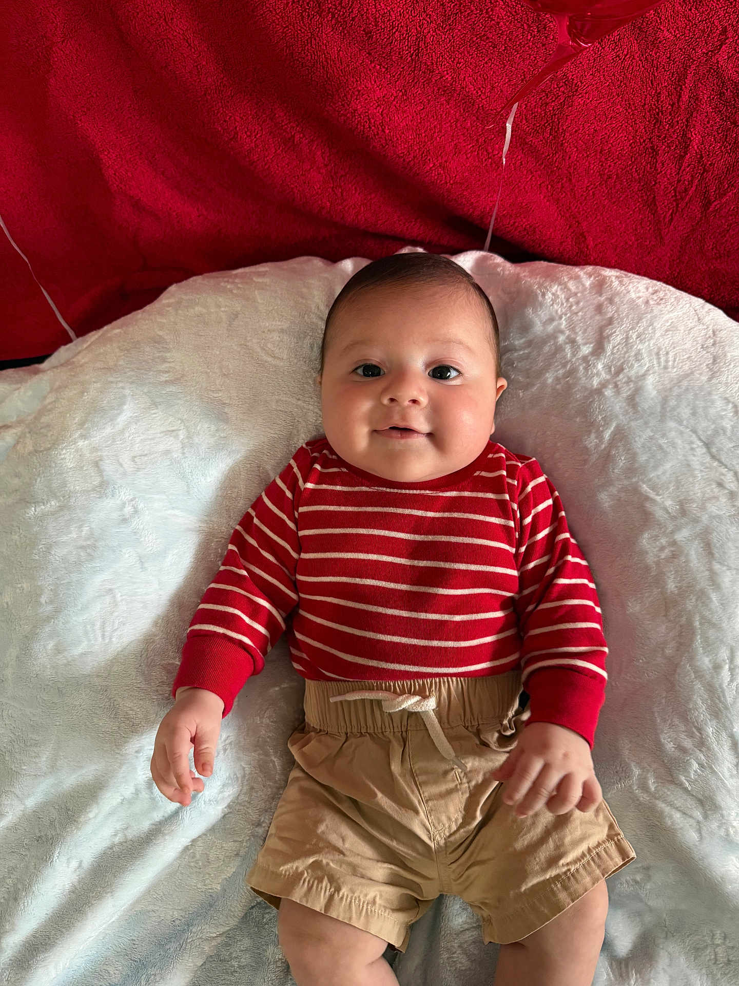Thiago is registered to the contest to win money with this photo: baby, infant, smile, red_striped_sweater, beige_shorts, blanket, soft_texture, face, child, cute, lying_down, indoors, skin, hand, head, eyebrows, cheeks, person, clothing, portrait