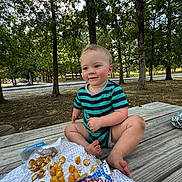 Noah joined the competition — help win amazing prizes! toddler, child, baby, striped_onesie, picnic_table, food, tater_tots, burger, outdoor, park, trees, daylight, grass, smiling, sitting, wood, nature, casual, playful, happy