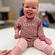 Ella joined the competition — help win amazing prizes! baby, toddler, child, tongue_out, pink_clothes, love_shirt, sitting, table, indoor, blurred_background, smiling, face, legs, shoes, hand, ring, bodysuit, portrait, bokeh, cute