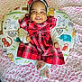 animal_print, baby, blanket, bow, child, cushion, cute, feet, happy, headband, indoors, infant, patterned_background, pink_flowers, plaid, portrait, red_dress, sitting, smiling, white_socks
