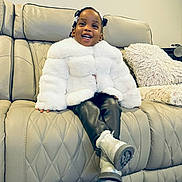 Camila is registered to the contest to win money with this photo: toddler, child, smile, white_coat, black_pants, silver_boots, leather_couch, cushion, indoor, happy, person, seated, hair_buns, fashion, portrait, cozy, home, furniture, cute, young_child
