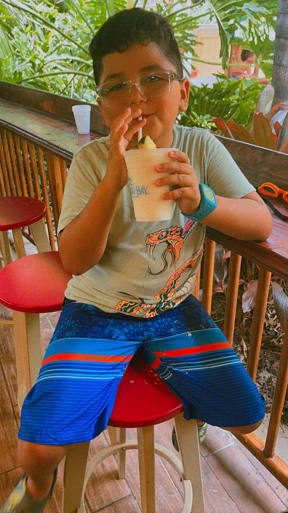 Xavi is registered to the contest to win money with this photo: barefoot, chair, child, electric_blue, eyewear, finger, fun, glasses, green, happy, human_leg, leisure, outdoor_furniture, person, plant, shorts, sitting, t_shirt, thigh, toddler