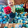 Tyden is registered to the contest to win money with this photo: toddler, child, ball_pit, building_blocks, red_block, green_block, smiling, play, indoor, dinosaur_mural, toy, person, adult, colorful, fun, curly_hair, casual_clothing, blue_shirt, gray_pants, wooden_floor