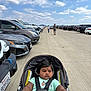 baby, stroller, parking_lot, car, sidewalk, child, person, outdoor, sky, cloud, road, vehicle, casual_clothing, seat_belt, head, face, walking, summer, sunny, people
