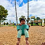 Ram is registered to the contest to win money with this photo: toddler, child, swing, playground, outdoor, park, wood_chips, green_swing, play_equipment, trees, cloudy_sky, grass, daytime, pants, barefoot, person, holding, expression, curious, recreation