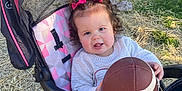 Sabrina is registered to the contest to win money with this photo: toddler, child, stroller, football, outdoor, grass, straw, pink_bows, curly_hair, white_sweater, pink_pants, toy, baby, seat, play, sunlight, park, nature, cute, expression