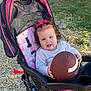 toddler, child, stroller, football, outdoor, grass, straw, pink_bows, curly_hair, white_sweater, pink_pants, toy, baby, seat, play, sunlight, park, nature, cute, expression