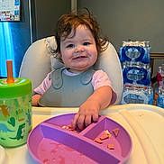 Sabrina is registered to the contest to win money with this photo: toddler, child, high_chair, plate, food, sippy_cup, dinosaur, curly_hair, smiling, kitchen, water_bottles, calendar, feeding, mealtime, plastic_plate, plastic_cup, baby_bib, happy, indoor, person
