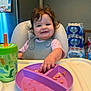 toddler, child, high_chair, plate, food, sippy_cup, dinosaur, curly_hair, smiling, kitchen, water_bottles, calendar, feeding, mealtime, plastic_plate, plastic_cup, baby_bib, happy, indoor, person