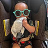 baby, car_seat, sunglasses, child, infant, cloth, seatbelt, colorful_clothing, indoor, cute, relaxed, chewing, sitting, safety_harness, head_support, black_fabric, toddler, accessory, funny_glasses, babywearing