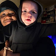 Orion joined the competition — help win amazing prizes! baby, adult, hoodie, indoor, smile, person, face, child, holding, warm_lighting, portrait, shelf, box, cozy, family, closeup, black_clothing, happy, casual, bonding