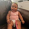baby, child, smiling, sitting, pink_clothing, hair_bows, diaper, pacifier, beaded_chain, leather_chair, cardboard_box, happy, cute, indoors, person, infant, plump_arms, plump_legs, casual, portrait